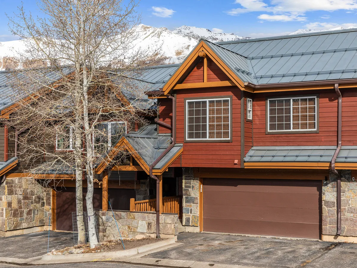 A welcoming exterior shot of a mountain lodge featuring a stone facade, wooden accents, sloped roof, and nearby trees. The lodge sits nestled among nature with the mountains as a dramatic backdrop, evoking a sense of arrival and community.