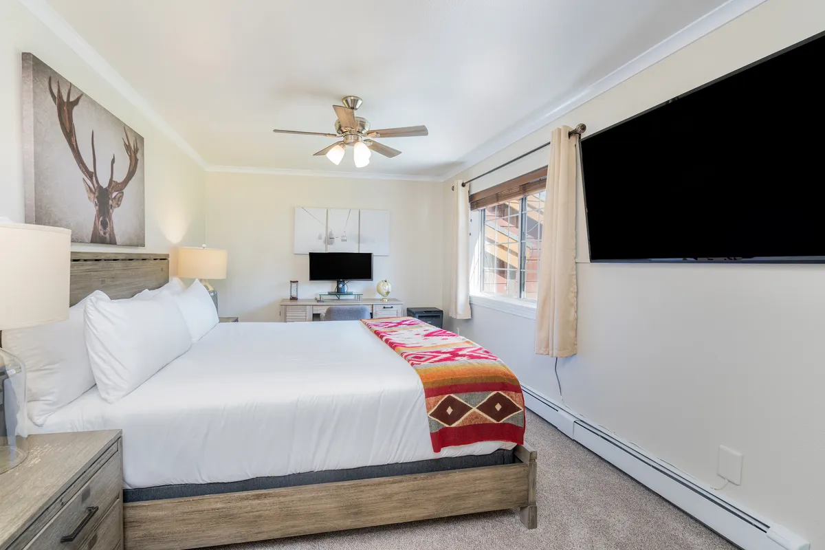 A serene bedroom featuring a plush bed adorned with a decorative blanket, a TV mounted across from the bed, a work desk, and soft natural light streaming in through a window. The overall ambiance is inviting and restful, ideal for relaxation after a day of adventure.