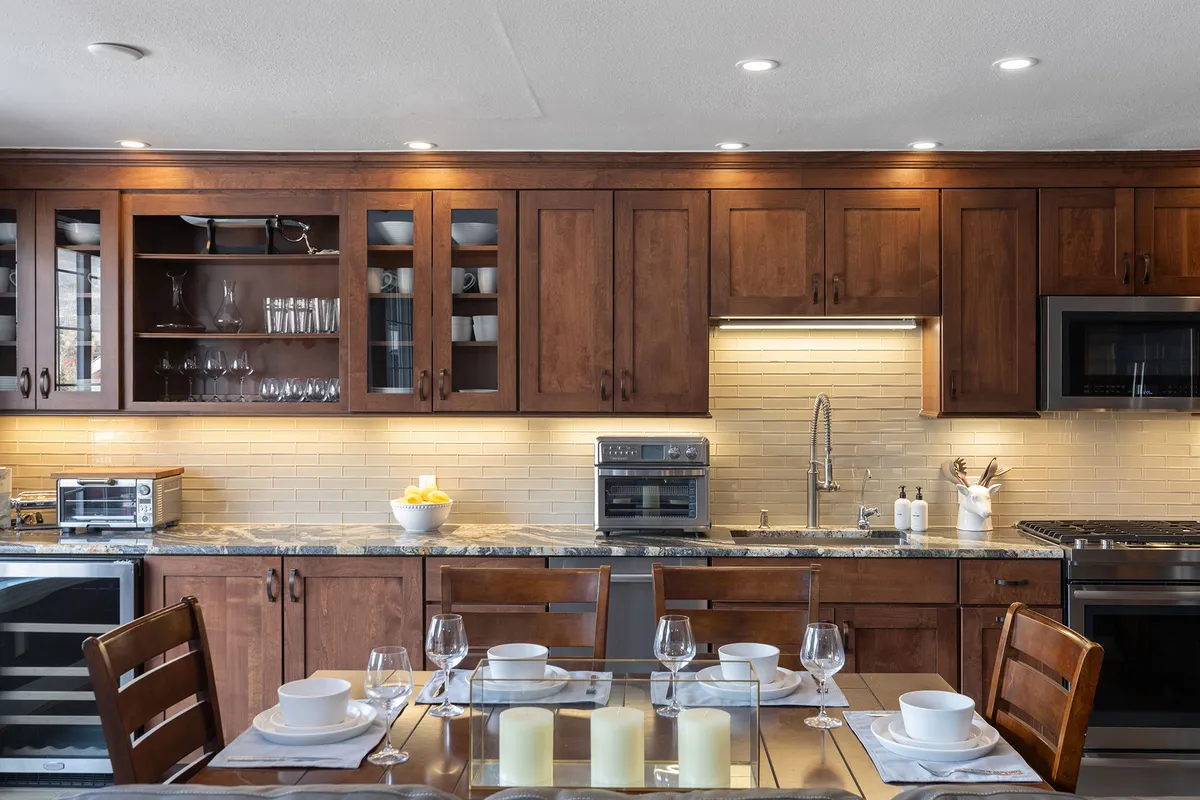 This image features a glass dining table set in a warm, inviting dining area. Wooden cabinets, kitchen appliances, and decorative bowls of fruit evoke the sense of a family-friendly space where meals and conversations are shared. The scene is well-lit and has a welcoming, homey atmosphere.