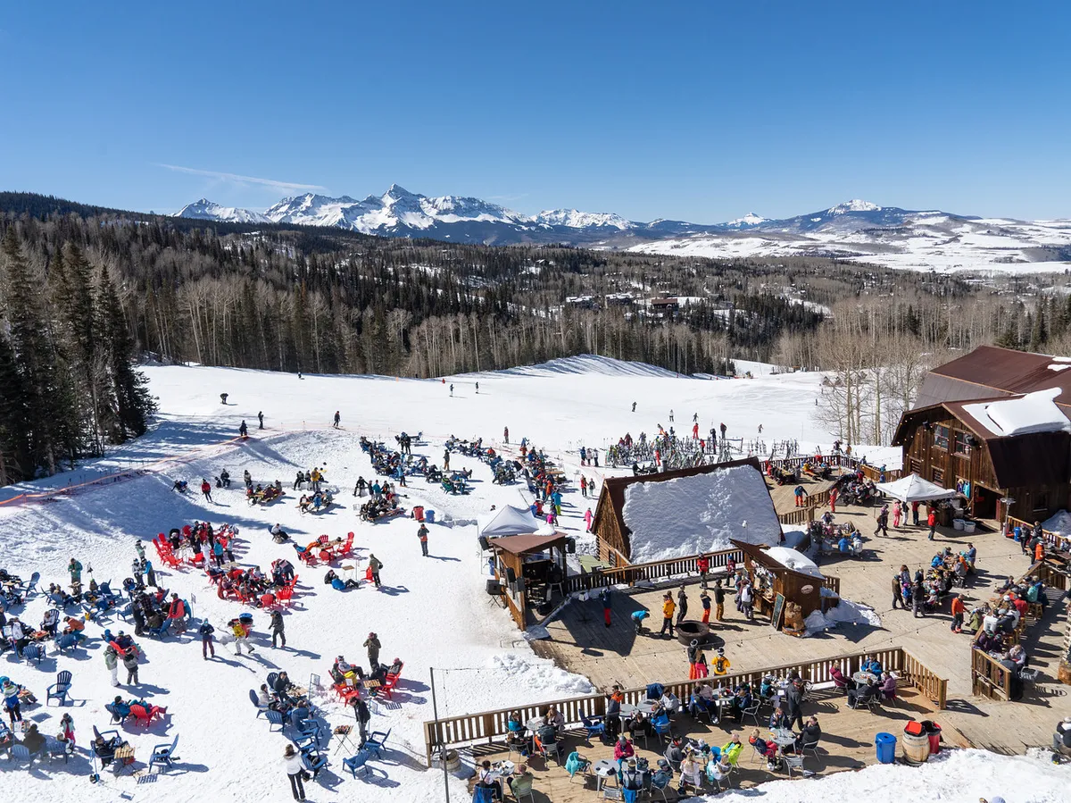 A vibrant winter scene featuring snow-covered slopes dotted with skiers in the distance. In the foreground, people are enjoying outdoor seating, suggesting a lively après-ski atmosphere against a stunning mountain backdrop.