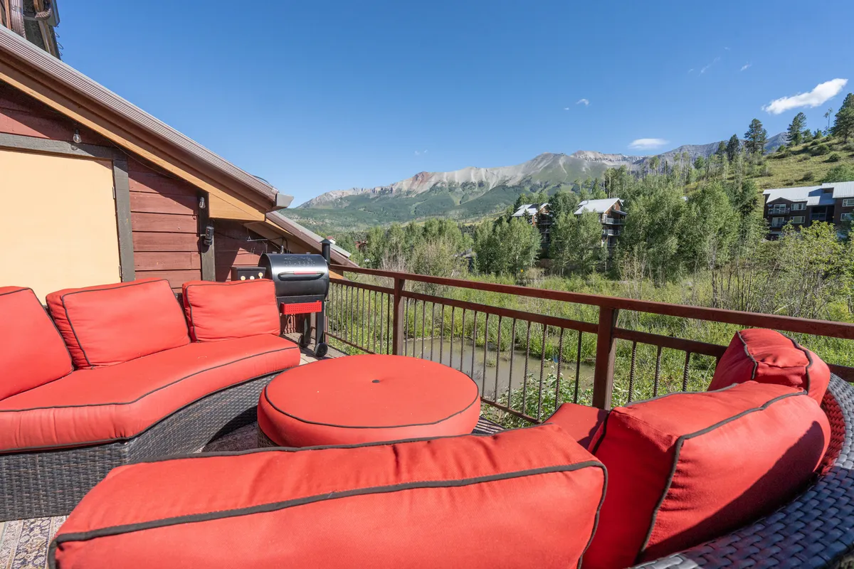 An exterior scene featuring comfortable red outdoor seating, a barbecue grill, verdant foliage, and panoramic mountain views. The arrangement suggests a welcoming spot for families to gather, relax, and socialize.