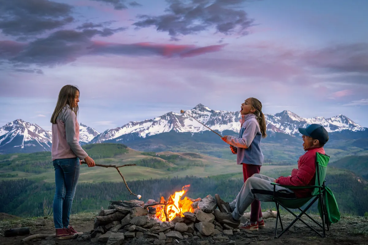 A joyful outdoor scene with children roasting marshmallows around a campfire, an adult relaxing in a chair, and a stunning mountain range illuminated by sunset in the background. The mood is warm, communal, and adventurous, encapsulating the spirit of summer family fun.