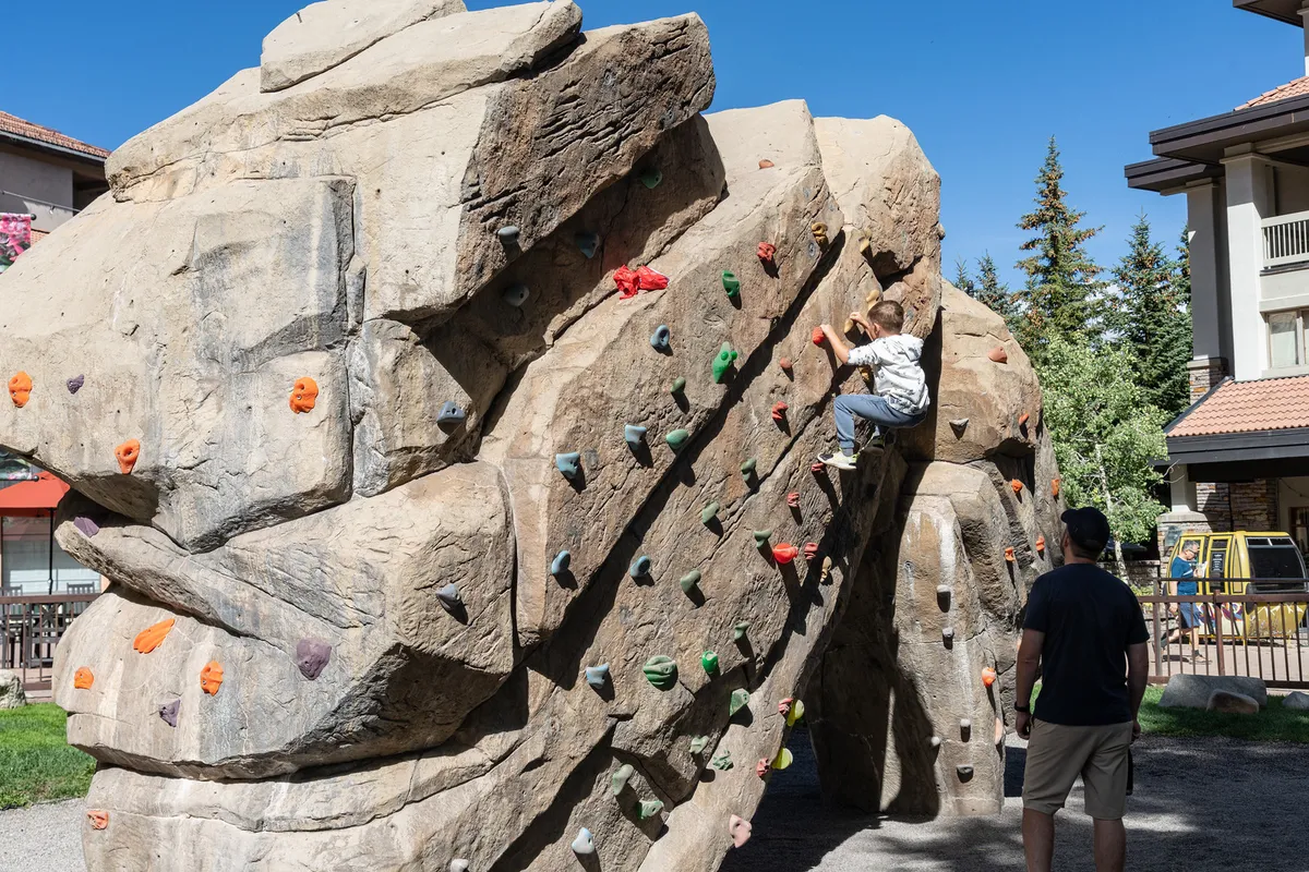 Outside, a child climbs a colorful climbing wall under the watchful eye of an adult. The scene is lively and playful, with clear blue skies and a sense of community and safety. It visually suggests nearby playground access and outdoor fun for kids.