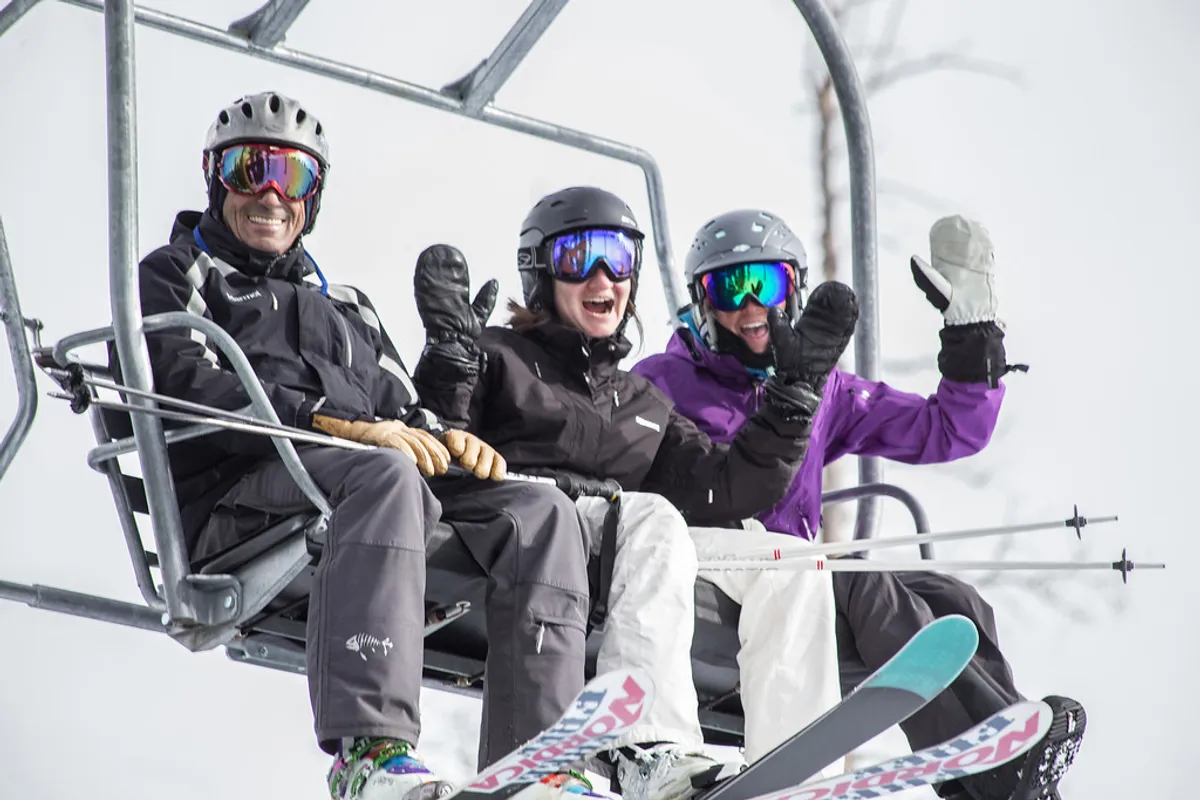 Three skiers are pictured riding a chairlift over a snowy landscape, with visible ski equipment and expansive white slopes beneath them. The mood is cheerful, emphasizing the excitement and camaraderie of a ski trip.
