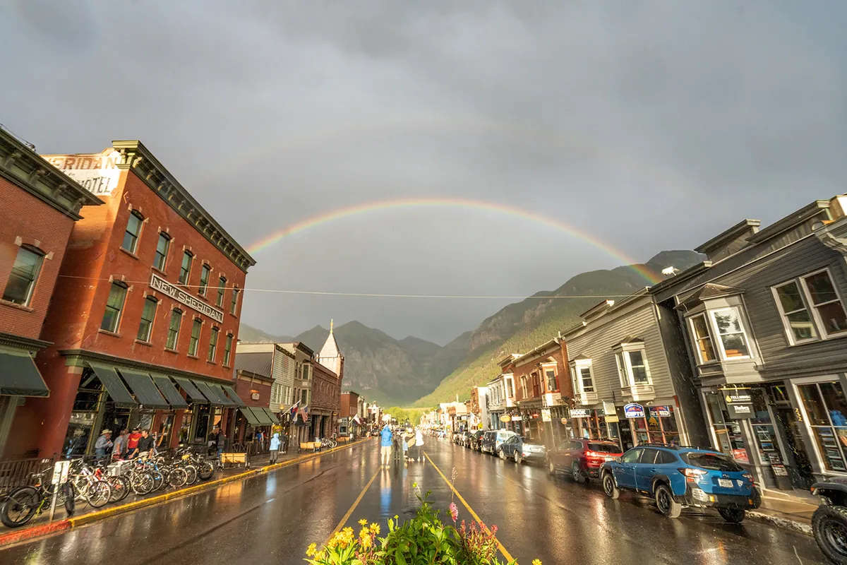 A cheerful town scene featuring a rainbow arched over historic buildings, wet streets, bicycles parked, and people strolling and exploring local shops. The setting is lively and welcoming, inviting outdoor activity and exploration.