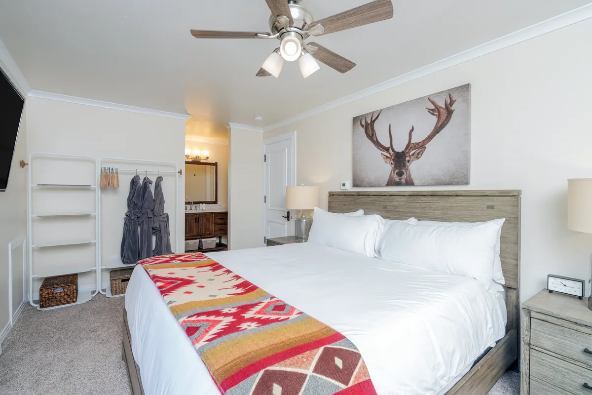 A cozy guest bedroom where a bed with a colorful blanket is the central feature, complemented by deer artwork on the wall, hanging robes by a vanity area, and abundant natural light. The scene feels bright, cheerful, and perfect for quiet moments.