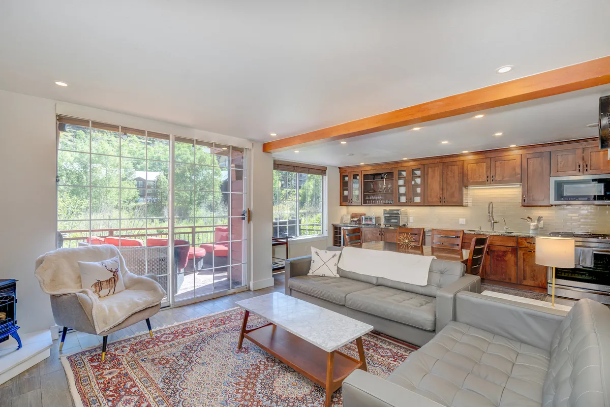 The image showcases a bright and inviting open-concept living area with floor-to-ceiling windows framing mountain sunrise views, a sleek modern kitchen, plush seating arranged around a decorative rug, and a warm, cozy atmosphere perfect for family gatherings or relaxing after a day on the slopes.