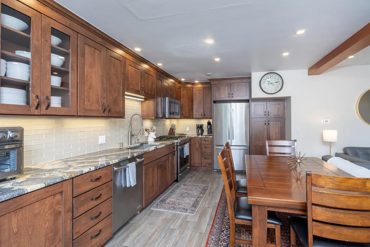 A wide view of a stylish open-concept space featuring a central kitchen island, adjacent dining table, modern appliances, and warm wooden cabinetry. The layout encourages social interaction, with the dining and kitchen areas seamlessly connected, inviting gatherings for meals, games, or casual conversation.
