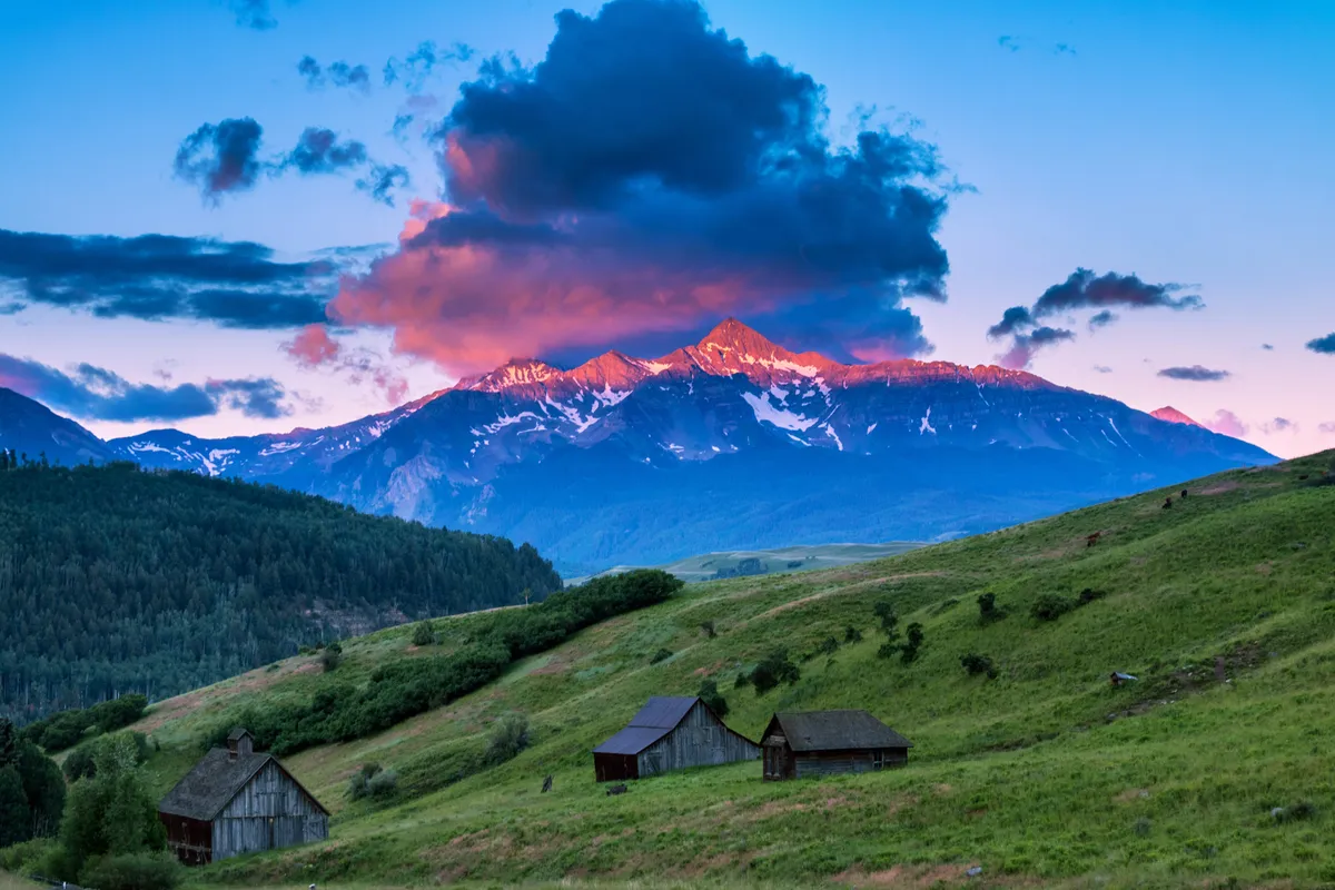 A wide, scenic exterior photograph capturing majestic mountains, clouds tinged with sunset colors, rustic cabins nestled within lush hills, and open spaces ideal for family walks or playtime.